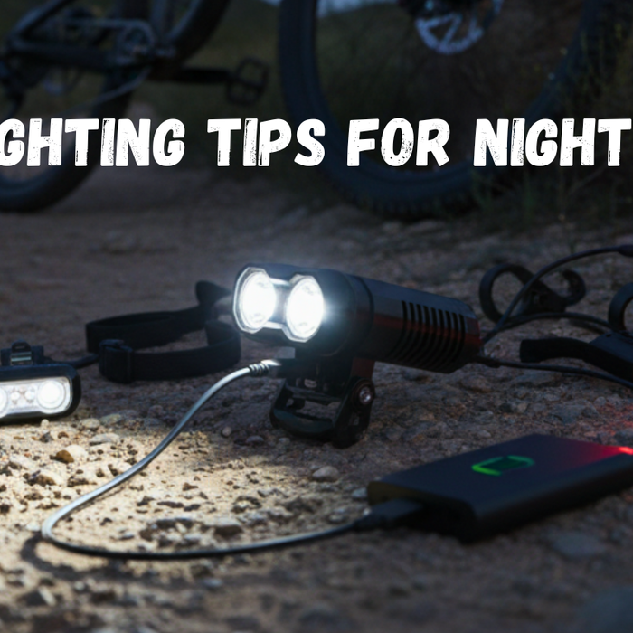 Mountain biker riding at night on a rugged trail with a bright front headlight illuminating the path and a glowing red rear light, surrounded by dark mountain scenery under a deep blue sky.