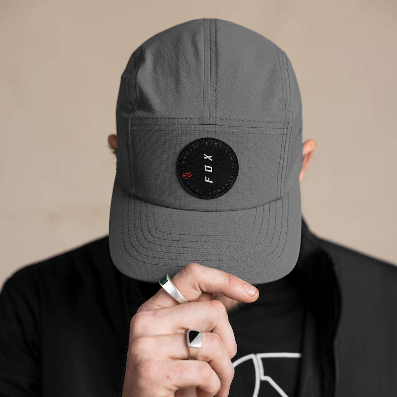 A grey hat with a circular graphic in the center, bearing the word "Fox" as a logo, is the main object in the picture. It is being held by an athlete's hand, who is looking down and partially visible in the image.